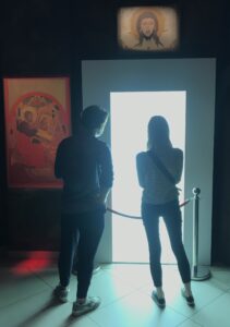 Dwie osoby stojące tyłem do widza na tle drzwi, skąd bije światło oślepiające.