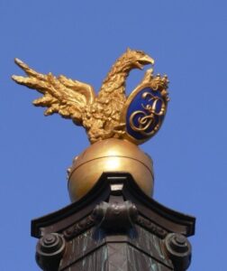 Złoty orzeł trzymający w szponach herb rodu Branickich. Tło herbu niebieskie w środku złote inicjały J K B.