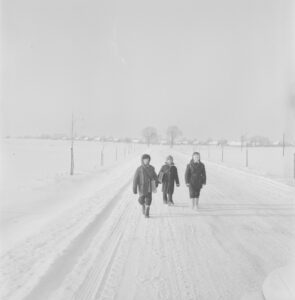 Trójka dzieci w zimowych ubraniach idzie przez drogę. Droga i okolice zaśnieżone.