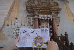 Na pierwszym planie dłonie przetrzymujące kartkę i lupę. W tle wnętrze synagogi w Tykocinie. Po lewej widoczna jest menora.