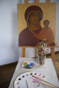 Na pierwszym planie pędzle i farbki do malowania. Na dalszym planie sztaluga z ikoną Matki Bożej i dzieciątkiem Jezus.