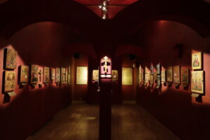 Sala wystawiennicza w Muzeum Ikon w Supraślu. Ściany koloru czerwonego, na ścianach wiszą ikony. Po środku na słupie zamontowany jest krzyż. 