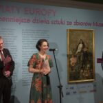 Dwóch mężczyzn i jedna kobieta po środku stoją na tle niebieskiej ściany z opisem wystawy. Kobieta przemawia do mikrofonu.