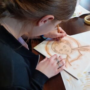 Dziewczyna szkicująca ikonę ołówkiem, w lewej ręce trzyma ołówek prawa przetrzymuje kartkę. 