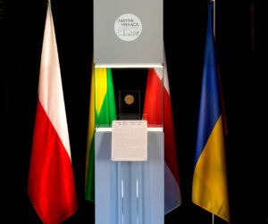 Na pierwszym planie szary kubik, przed nim stojak z informacją, w kubiku medal ustawiony na ekspozytorze z płyty plexi, od lewej flagi Polski, Litwy, Białorusi i Ukrainy.