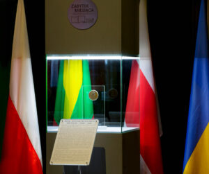 Na pierwszym planie szary kubik, przed nim stojak z informacją, w kubiku medal ustawiony na ekspozytorze z płyty plexi, od lewej flagi Polski, Litwy, Białorusi i Ukrainy.