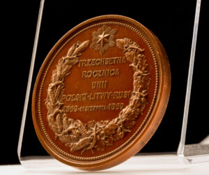 Na pierwszym planie widać zabytkowy medal pamiątkowy ustawiony na ekspozytorze z płyty plexi.