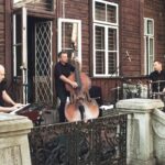 Trzech muzyków z instrumentami grająca przed drewnianym budynkiem